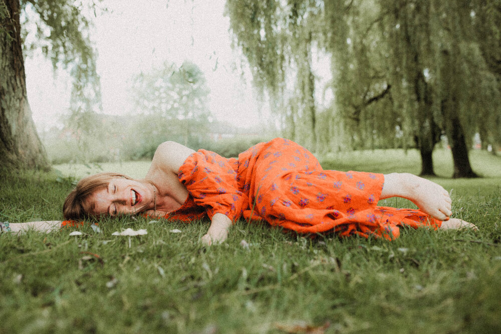 A woman lying on the grass, happily laughing, backed by sweeping willow trees.