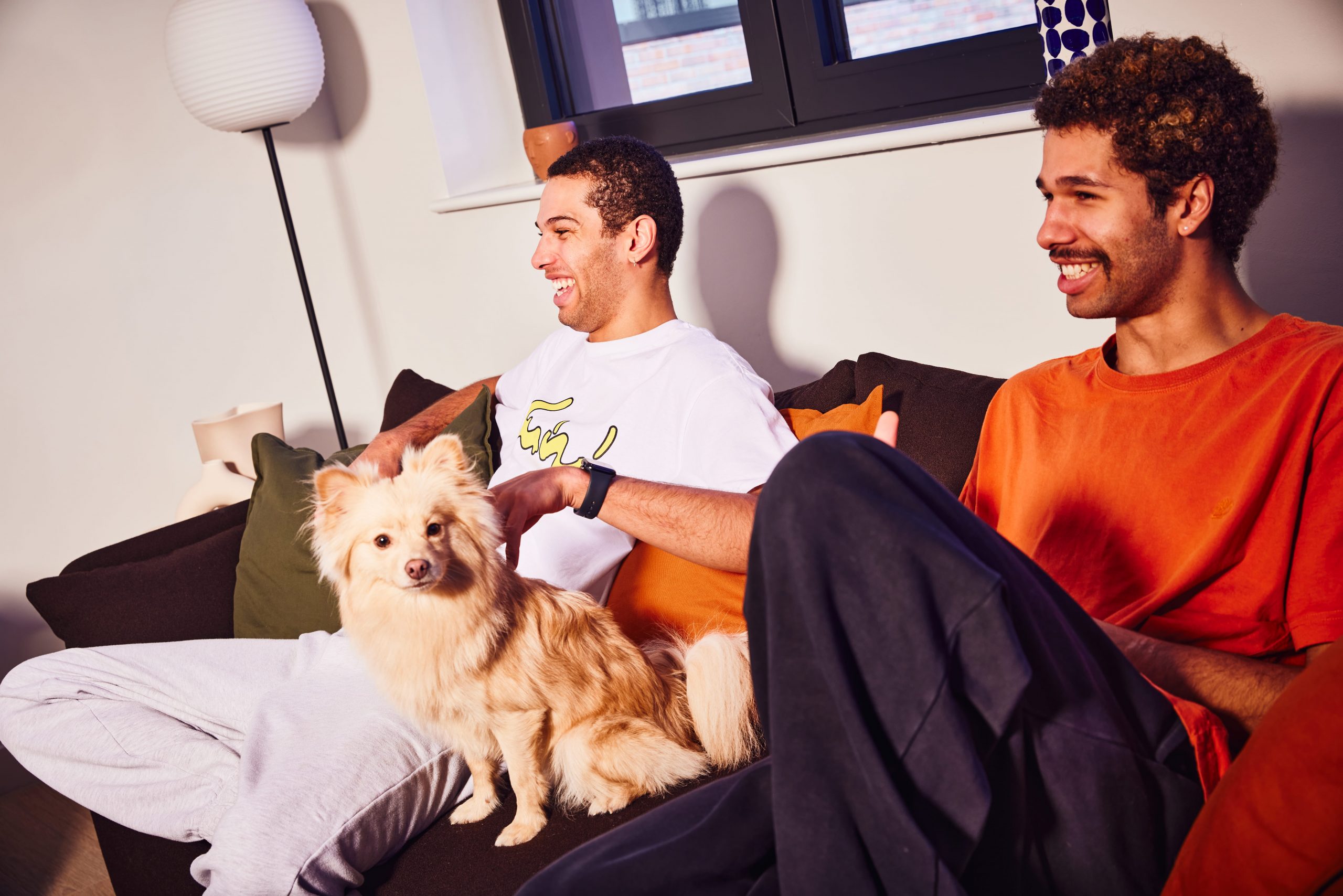 Two young people and a dog sit on a sofa, and laugh at a tv show.